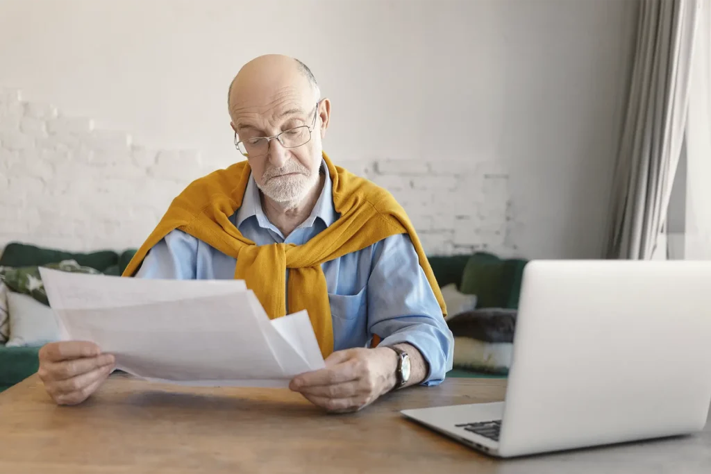 Older man with glasses in a blue shirt, mustard sweater draped over his shoulders, reading papers at a wooden table with a laptop nearby.